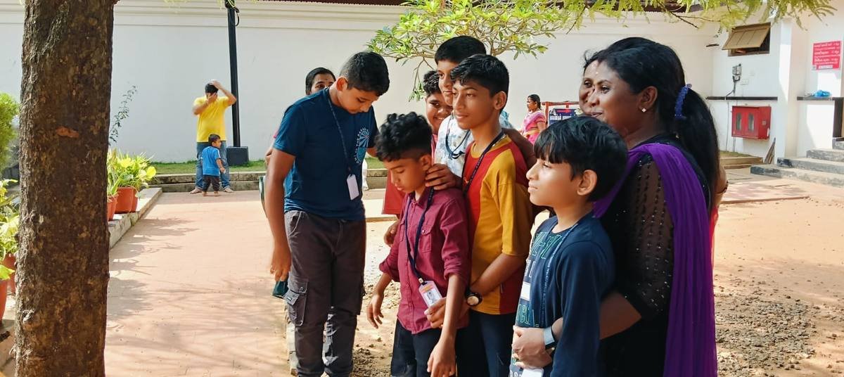 Pupils waiting in the shade of a tree, a teacher standing with them.